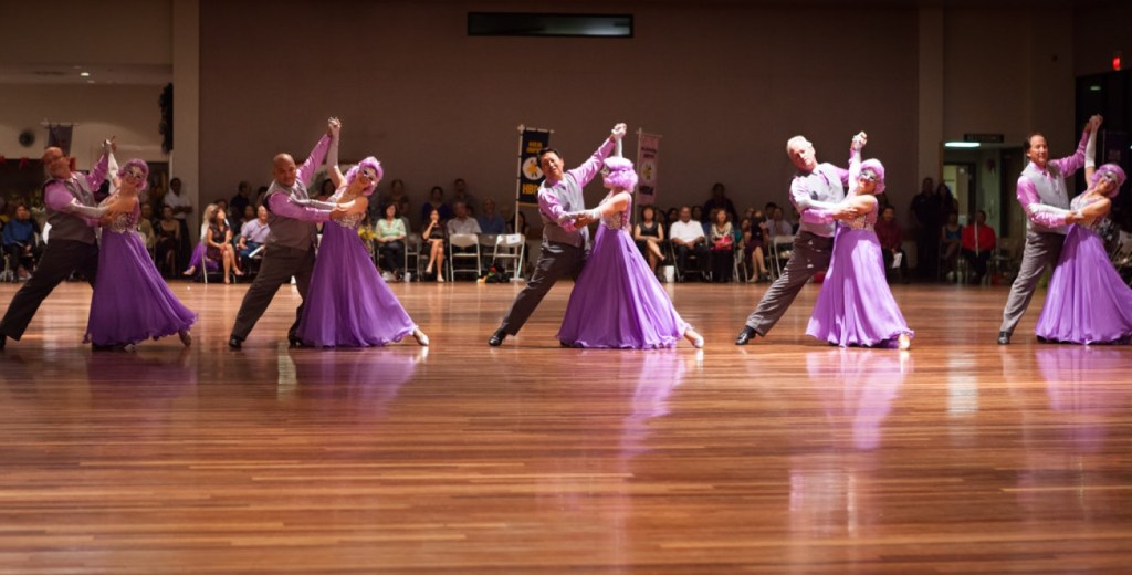 Five HBDA couples in purple gowns dancing a waltz across a wooden ballroom floor
